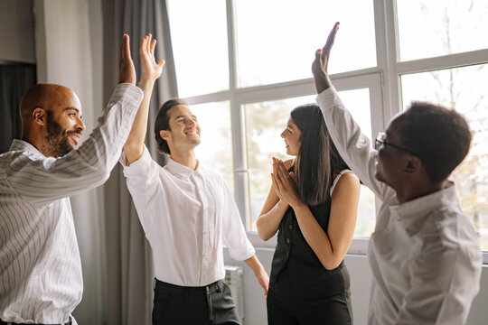 Four diverse office workers high-fiving and celebrating success indoors in light office
