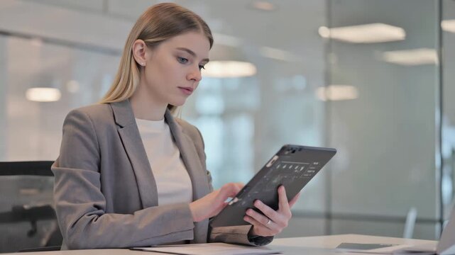 Young European female fintech developer engaged in reviewing digital payment interface on tablet while seated at modern office desk with bright lighting