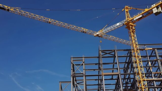Large high-rise building construction with steel framework extending upward against clear blue sky, showcasing crane lifting materials for structural assembly