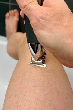 Man trimming his hairy legs with an electric trimmer in a bathtub, modern male grooming routine at home, intimate bathroom lifestyle scene.