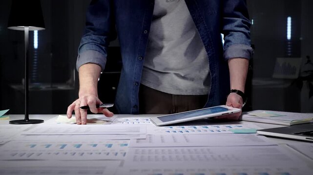 Entrepreneur standing at a desk in a dark office, studying financial reports and charts by lamp light while working on a laptop, focused on strategic planning and data analysis
