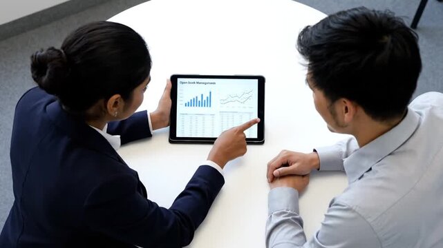 Two professionals, a woman and a man, analyze digital data and graphs on a tablet during a meeting, pointing and discussing various charts and trends.