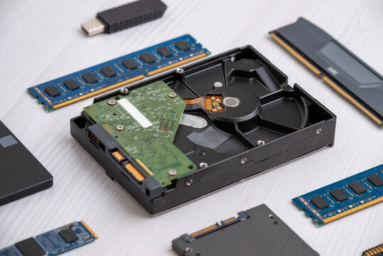 Opened hard disk drive with visible platter and circuit board surrounded by RAM modules and SSD on wooden desk