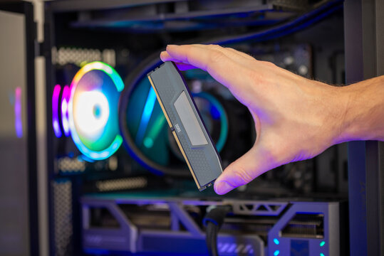 Hand holding DDR5 RAM module in front of gaming computer with RGB lighting, memory upgrade for high performance system
