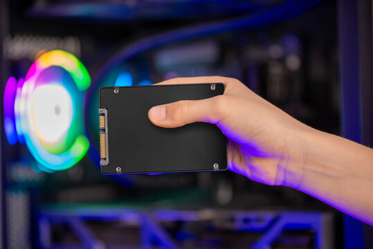 Hand holding SATA solid state drive in front of gaming computer with RGB lighting, storage upgrade and performance concept