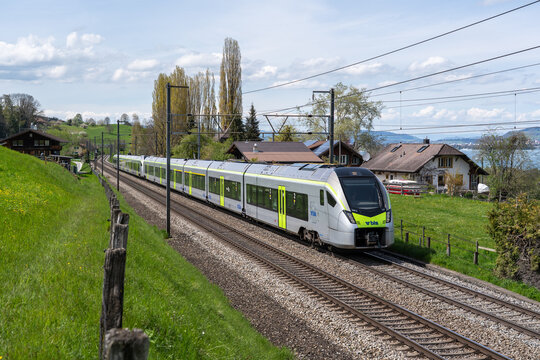 Spiez, Switzerland - April 27, 2023: Modern BLS passenger train near Spiez on a sunny day, set against beautiful Swiss landscape with mountains, green fields, and blue sky.