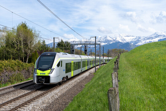 Spiez, Switzerland - April 27, 2023: Modern BLS passenger train near Spiez on a sunny day, set against beautiful Swiss landscape with mountains, green fields, and blue sky.