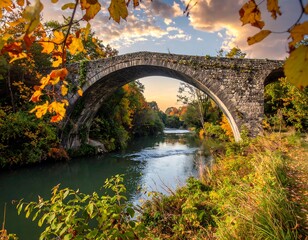 Obraz premium An aged stone bridge arcs over a river, framed by autumn foliage under a partly cloudy sky at golden hour