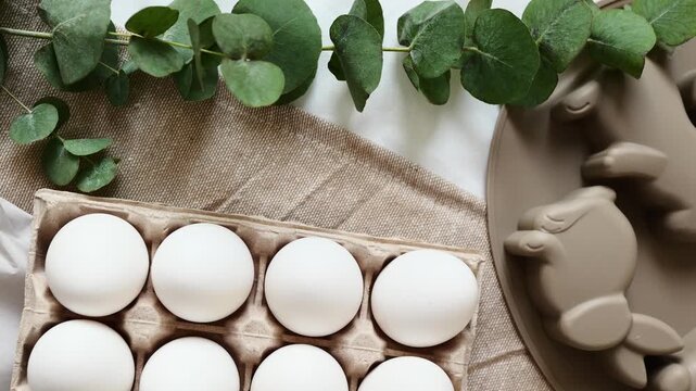 A bunny shaped dish holds eggs next to an egg carton on a cloth. Greenery decorates the setting.