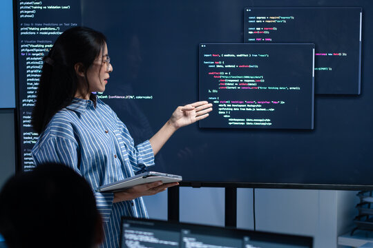 Asian female software engineer presenting source code on large screens in a modern tech office, explaining solutions in a team meeting. Concept for coding, code review, agile workflow.