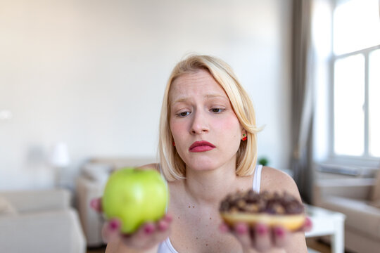 Decision making for healthy lifestyle. Worried woman holding green apple and chocolate donut, Dieting and weight loss concept. Attractive blonde girl looking at unhealthy food with pained face.