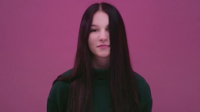 Portrait: young smiling Caucasian woman teen with long naturally black hair. White teenager brunette pretty girl, with green pullover smiles warmly at the camera. Pink background