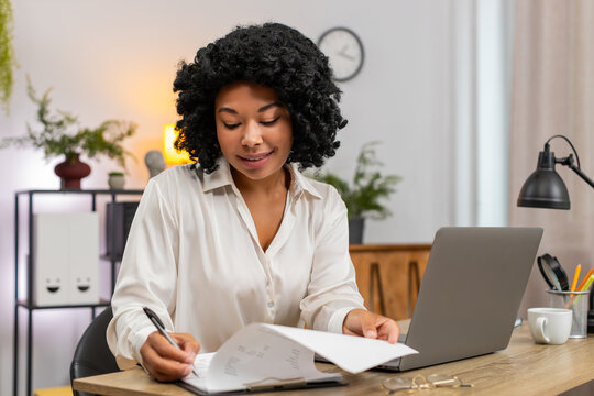 African American businesswoman at home office matches documents to laptop data smiling satisfied. Freelancer girl at table ticks paper list with pen very pleased by accurate results feeling organized