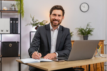 Obraz premium Middle-aged businessman at home office watches webinar on laptop wearing earphones takes notes and smiles. Freelancer guy at table snaps smartphone photo of screen and taps key to change slide today.
