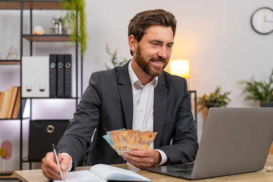 Middle-aged businessman at home office counts cash euros and enters amounts on laptop for daily budget well. Freelancer guy at table writes notes in notebook adds sums and plans expenses and savings.