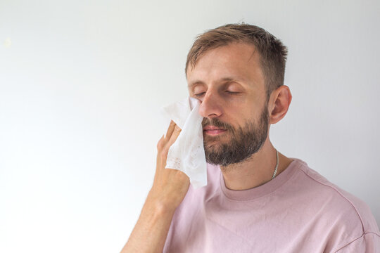 Adult man with short brown hair and trimmed beard wiping face with tissue after facial mask or scrub, eyes closed, light pink t shirt on white background, concept skincare routine, self care, 