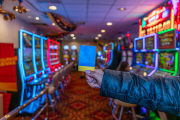 Fototapeta premium Blue and yellow card held by a hand against a colorful casino background with neon lights and slot machines, creating a modern entertainment setting.
