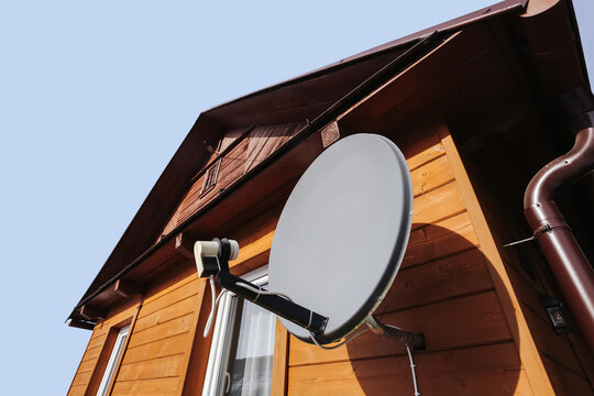 Satellite dish mounted on wooden house facade. Home television antenna receiver on residential building exterior. Modern communication technology installed on rooftop for signal reception.