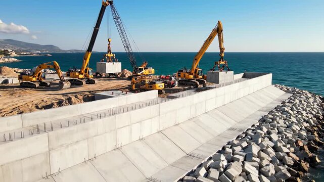 Construction site with yellow excavators lifting concrete blocks onto a coastal structure, showcasing the progression of building a seawall along the shoreline