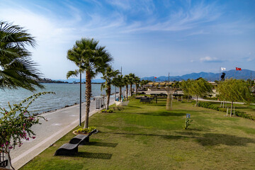 Obraz premium Scenic seafront promenade with palm trees, green lawn and sun loungers in Turkey. Beautiful mediterranean park landscape under a bright sky. Summer resort atmosphere in Fethiye.