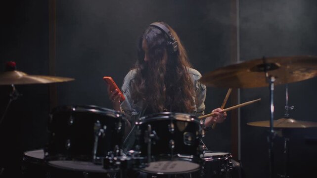 Woman drummer use cellphone in recording rehearsal studio, surrounded by drums, cymbals. Girl musician texting or typing messages, social network. Female with long curly hair makes a post on Internet