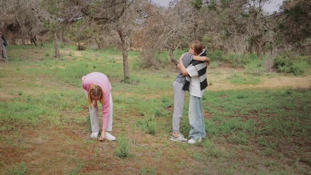 Group of three young women in casual clothing interact in a green outdoor setting, with one woman hugging another while a third woman observes and picks something from the ground