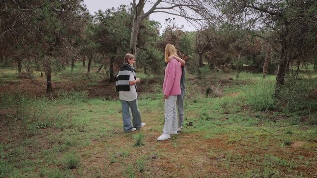 Group of children in a forest interact playfully, with one child hugging another while a third child observes nearby, surrounded by trees and greenery in a natural outdoor setting