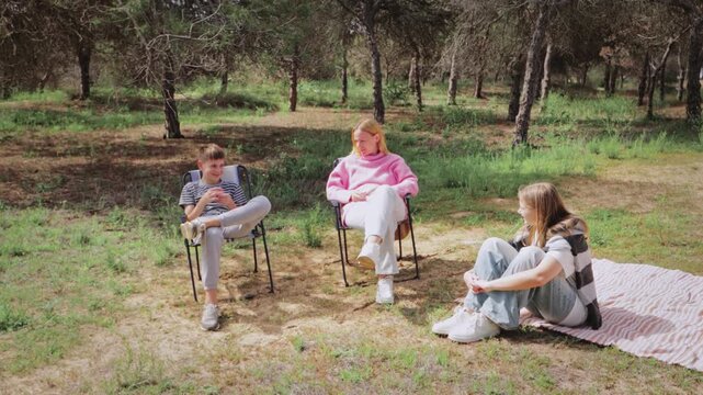 Group of three young friends relax outdoors in a forest clearing, with two seated in folding chairs and one on a blanket, enjoying a sunny day surrounded by trees and greenery