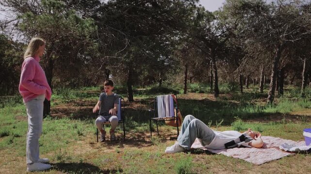Group of young friends interact in a sunny forest clearing, with one seated on a chair, another lying on a blanket, and a third standing nearby, enjoying a relaxed outdoor moment together