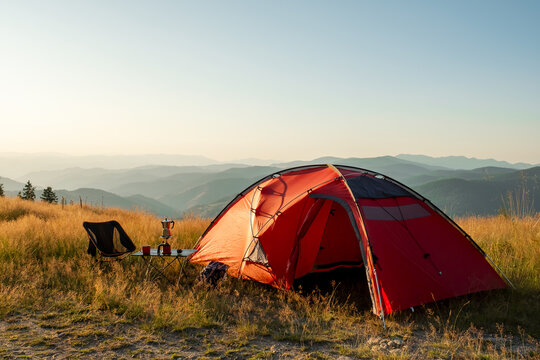 Cozy campsite mountain meadow golden hour. Red tourist tent pitched field. Small table with espresso maker metal mugs. Beautiful landscape vista. Peaceful trekking vacation adventure nomadic life