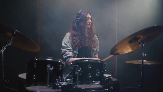 Beautiful drummer girl with long hair is tapping rhythm and drum part in rehearsal room. Young female with headphones performs solo on stage, kit. Sound checking equipment before a rock concert