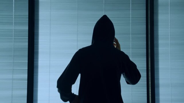 Back view shot of hacker with black hoodie clothes talking on mobile phone. Holding smartphone mysterious man standing by closed office or apartment window at night with shut blinds or shutters