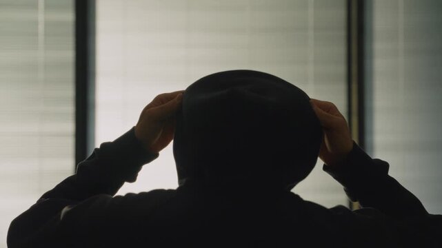Mysterious person put on black hoodie cap standing by office or apartment window.  Anonymous hacker man or spy wearing black clothes. Suspicious standing indoor. Criminal cyber intelligence activity