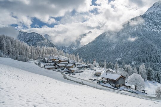 Snow-covered Bschlabs in Tyrol, Austria, nestled in the Lechtal Alps. Quiet alpine village in winter with deep snow, dramatic mountains, and soft sunlight breaking through mixed clouds.