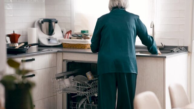 Senior woman in green pajamas unloading the dishwasher in a modern kitchen. Domestic routine of an elderly person doing daily household chores