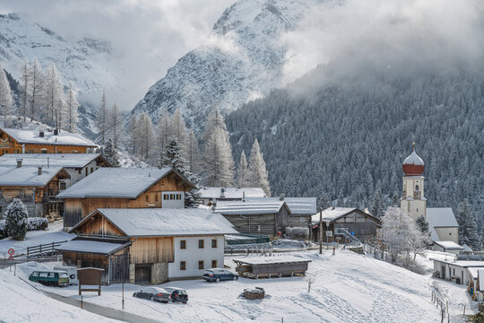 Snow-covered Bschlabs in Tyrol, Austria, nestled in the Lechtal Alps. Quiet alpine village in winter with deep snow, dramatic mountains, and soft sunlight breaking through mixed clouds.