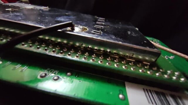Macro view of a green PCB featuring a metal EMI shield, integrated circuits, and a precision soldered wire connection on a complex electronic motherboard.