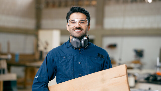 Enthusiastic carpenter wearing safety goggles and ear defenders while holding wood, standing in his woodworking workshop