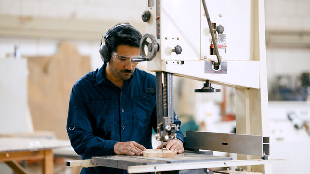 Woodworker operating a bandsaw to cut timber in a workshop while wearing protective safety goggles and ear protection