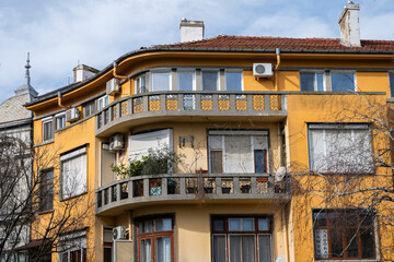 an ochre-colored apartment building featuring a rounded corner with matching curved concrete balconies, set against a cloudy sky and framed by bare tree branches.