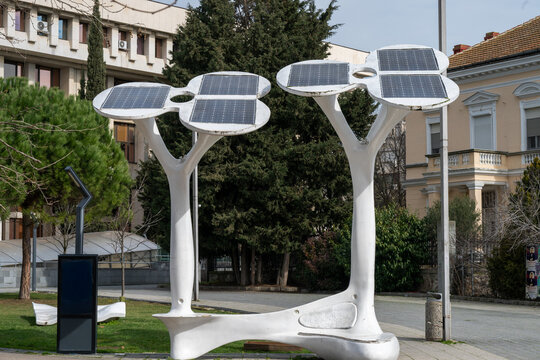 a modern, white, tree-shaped public seating structure featuring integrated benches at its base and a canopy made of multiple rectangular solar panels, set on a paved urban sidewalk near trees and back