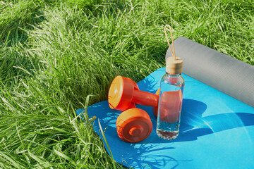 Glass bottle of water, dumbbells and blue yoga mat on green lawn Close-up