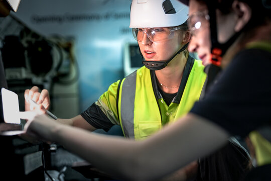 Two CNC programmers collaborate on the factory floor, using a laptop to upload and edit G-code for a 5-axis milling machine to ensure precision manufacturing of a complex component.