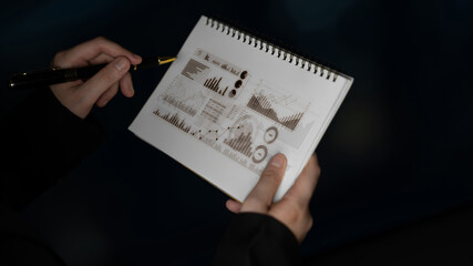 Close-up of hands holding a sketchbook featuring charts and graphs, illustrating business analysis and data visualization in a dark, professional setting. Scalp
