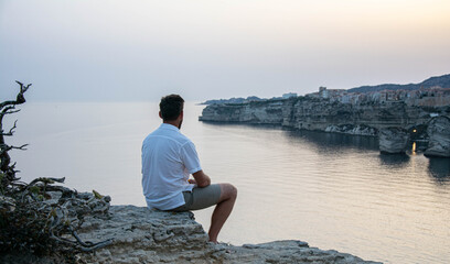 Man sitting on cliff overlooking calm sea at sunset