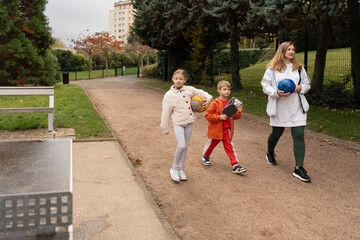 Fototapeta premium Mother and kids walking park path carrying balls and paddles
