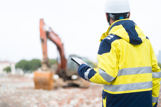Construction engineer in high-visibility jacket holding tablet while observing excavator operation on site