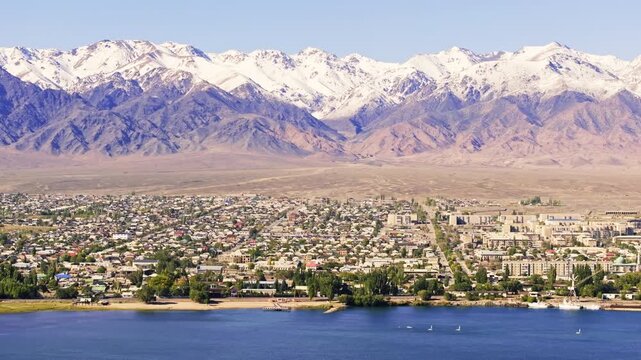 Aerial view captures majestic mountain range adjacent to Balykchy city. Sailboats glide across tranquil Issyk-Kul lake under clear daytime sky, creating picturesque scenery.