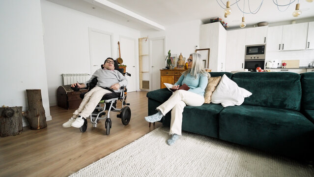 Man living with developmental condition relaxing in his electric wheelchair next to a woman sitting on the couch at home