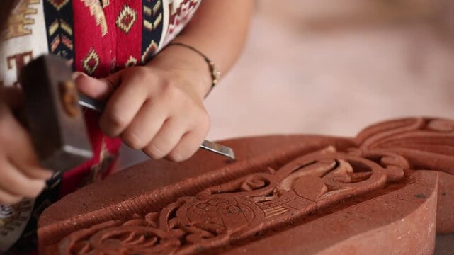 A skilled female artisan in colorful traditional attire uses a small hammer and chisel to meticulously carve an elegant floral pattern into a piece of red terracotta clay.

